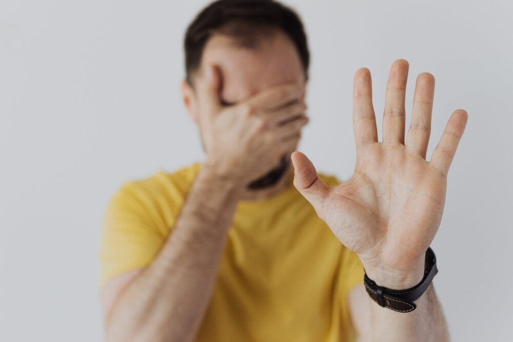 man in yellow shirt covering his face with hand and showing stop sign gesture